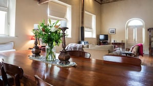 A dining area with a wooden table, chairs, and a vase with flowers. There is a television in the background and a window with a view of the outside. - Clare Valley Heritage Retreat (Clare)