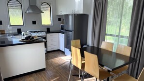 A modern kitchen with a central island, dining table, and chairs, with large windows and a view of greenery. - Barwon Valley Lodge (Belmont)