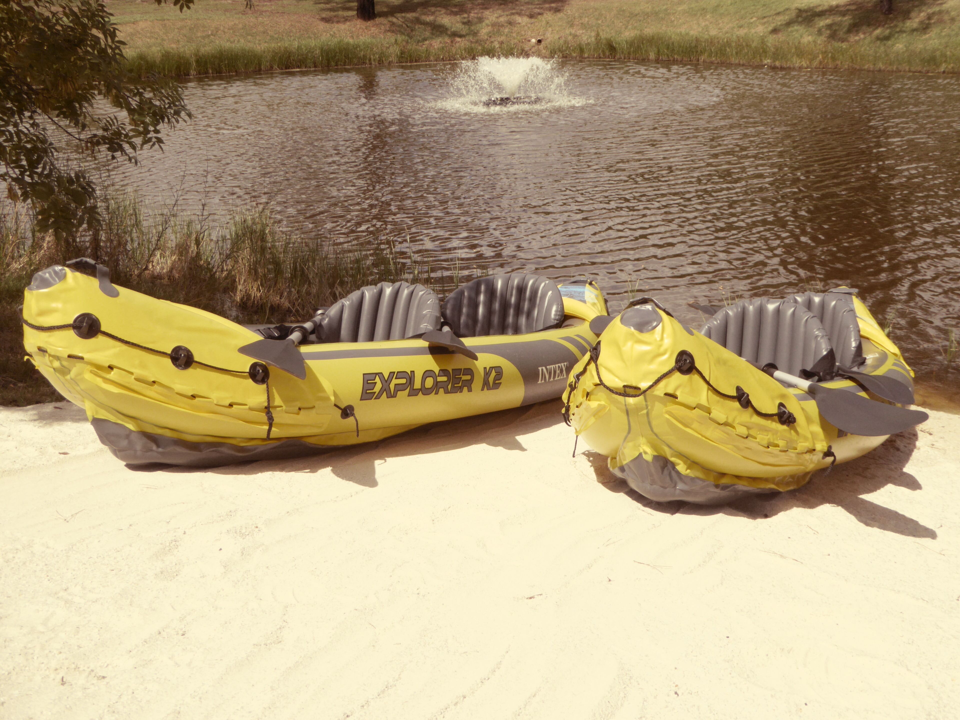 Dos kayaks inflables amarillos con la etiqueta 'EXPLORER K2' sobre una orilla arenosa junto a un cuerpo de agua.