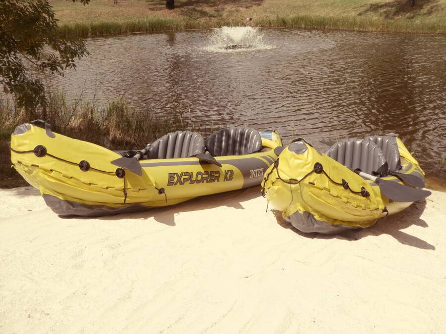 Dos kayaks inflables amarillos con la etiqueta 'EXPLORER K2' sobre una orilla arenosa junto a un cuerpo de agua.
