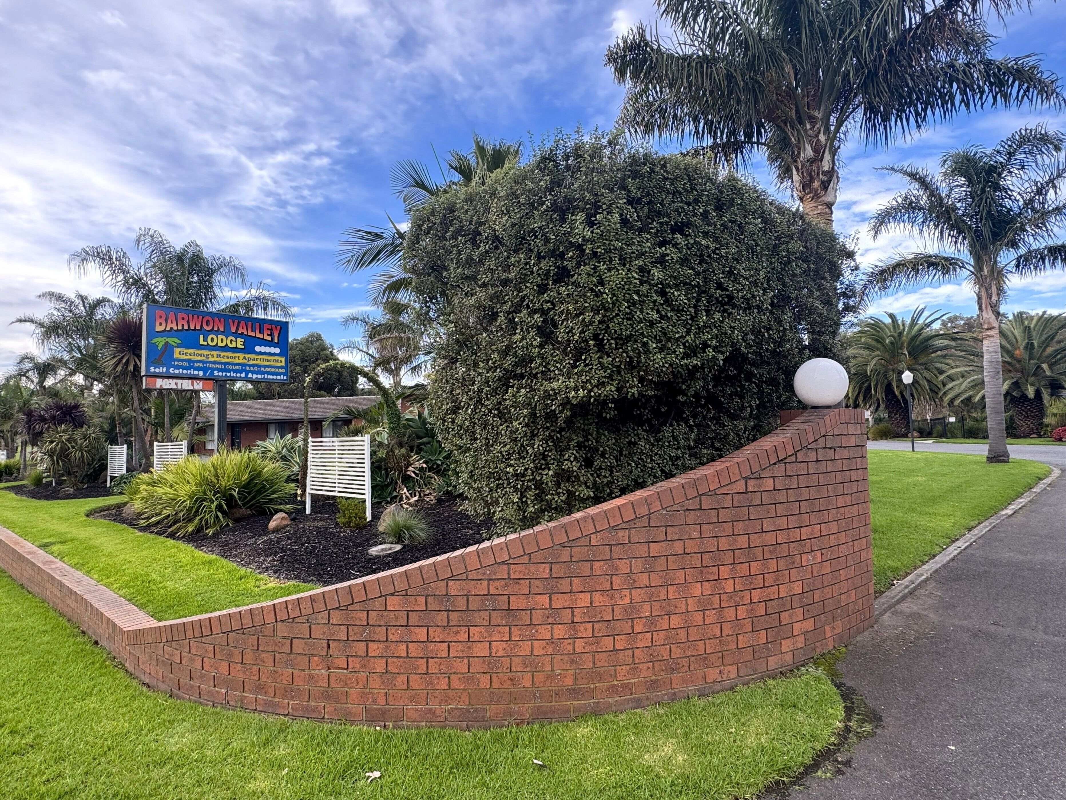 Letrero del Barwon Valley Lodge, un área ajardinada con un muro de contención de ladrillo y palmeras.