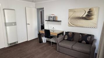 A room with a desk, a chair, a sofa, a radiator, and a wall-mounted shelf with a speaker.