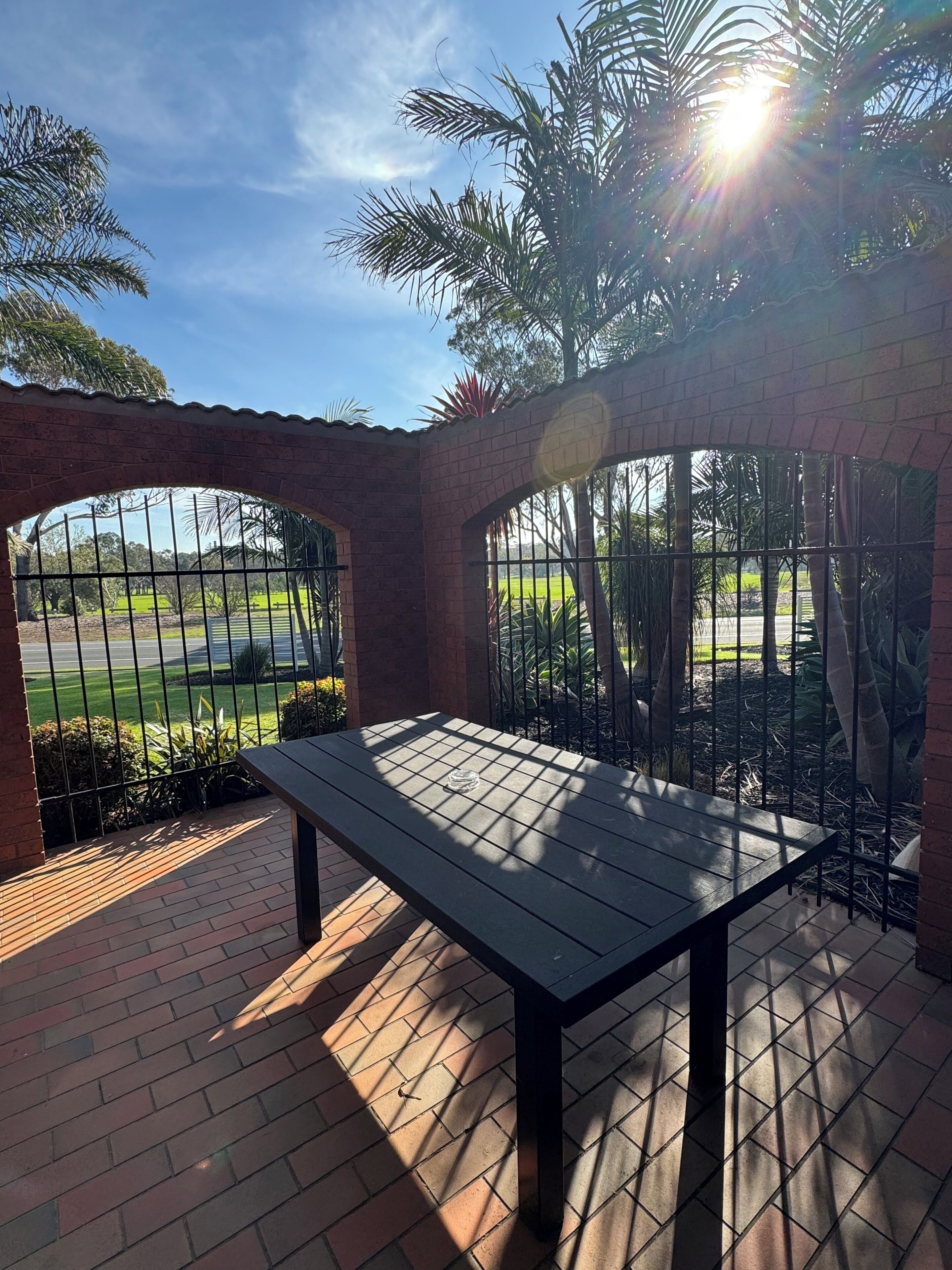 A patio with a table, brick flooring, and a view through a metal gate.