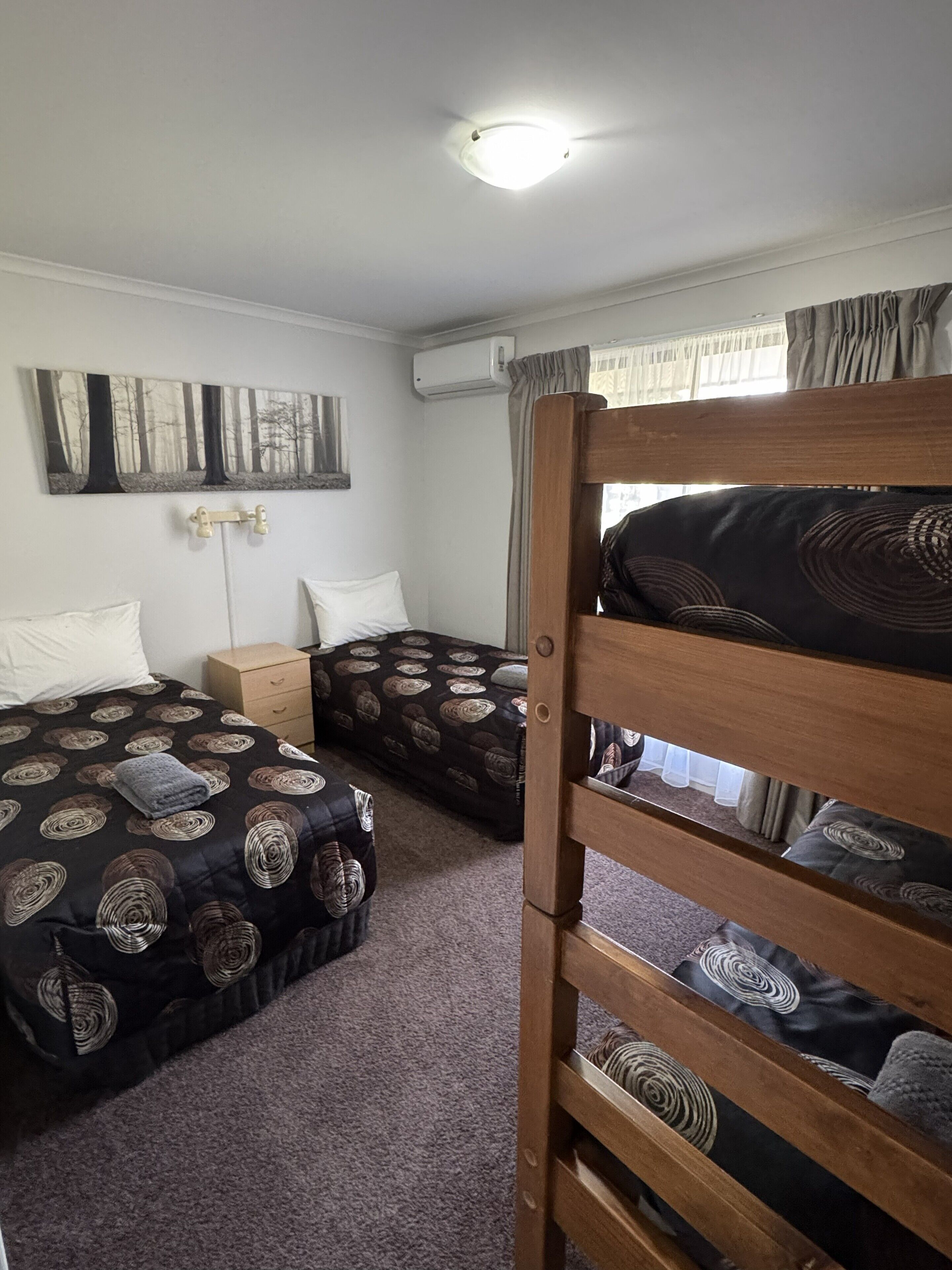 A room with two beds, a wooden bunk bed, a wall-mounted air conditioner, a small wooden nightstand, and a painting of trees.