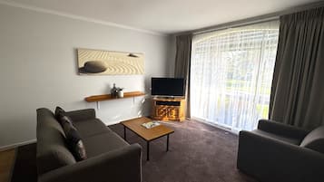 Una sala de estar con un sofá, una mesa de centro, un televisor sobre un mueble de madera y un ventanal con cortinas.