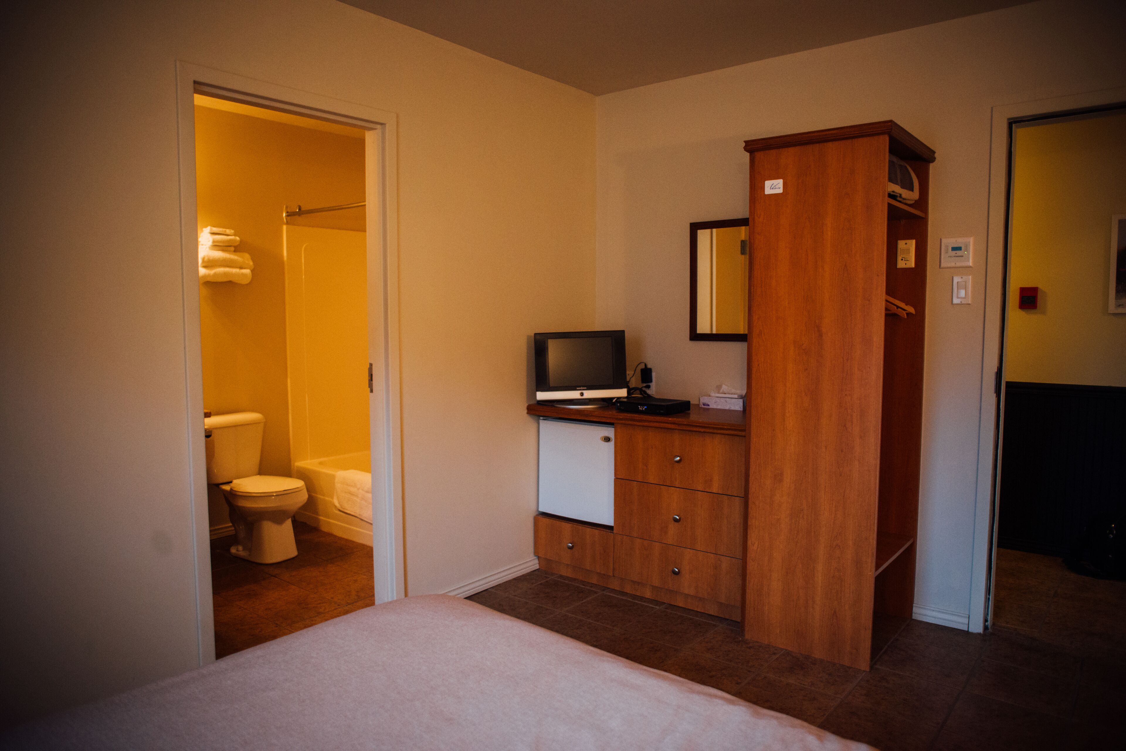 Une chambre d’hôtel avec un lit, une armoire en bois, un téléviseur et une salle de bain visible à travers une porte ouverte.