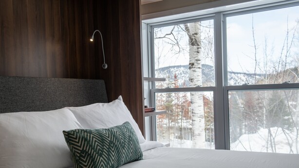 A modern bedroom with a large window, a wooden headboard, and a green patterned pillow. - Maison de Vébron (L'Anse-Saint-Jean)