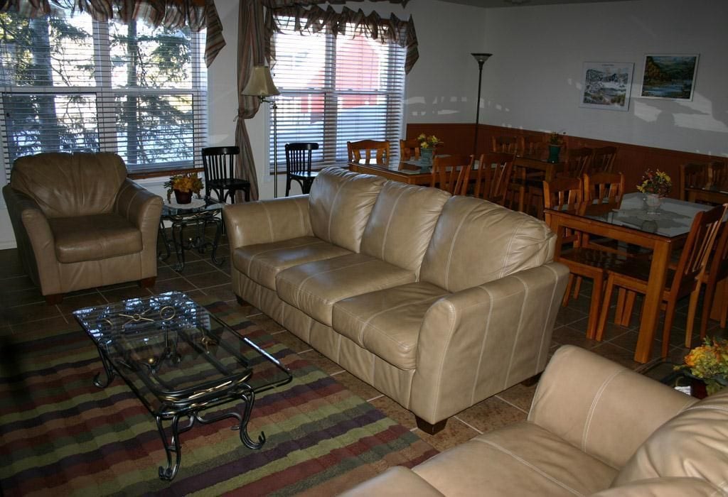 A living room with a beige leather sofa, a glass coffee table, and a striped rug. There are wooden chairs and a dining table with chairs.