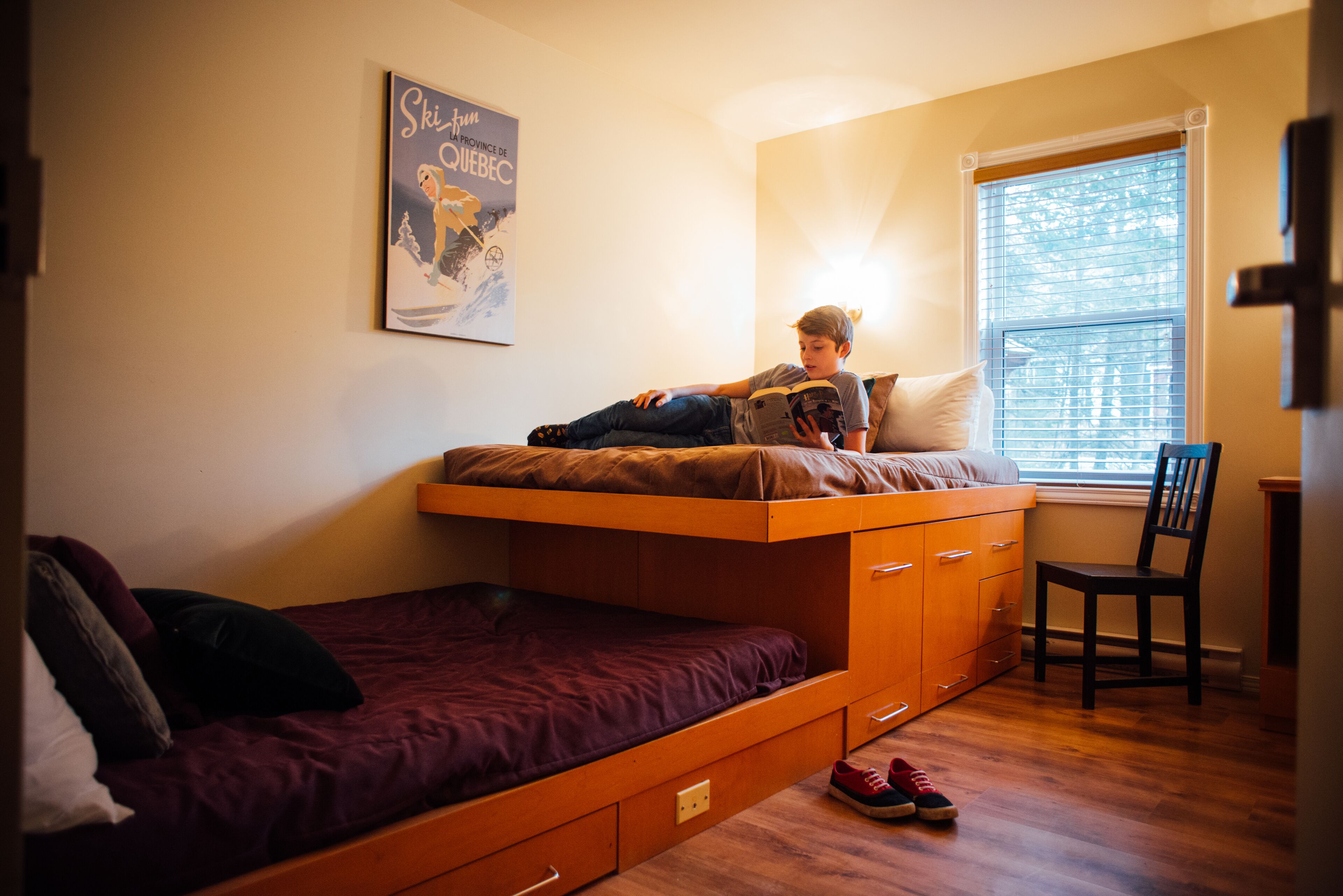 A bunk bed with a lower bed and a single bed, a poster on the wall, a chair, and a window with blinds.