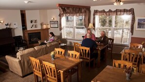 A cozy living room with a fireplace, a dining area with wooden furniture, and large windows with curtains. - Maison de Vébron (L'Anse-Saint-Jean)