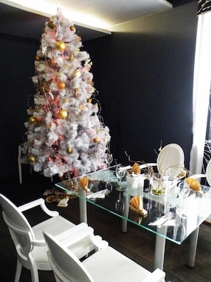 Un coin repas avec une table en verre, des chaises blanches et un sapin de Noël décoré d’ornements dorés.