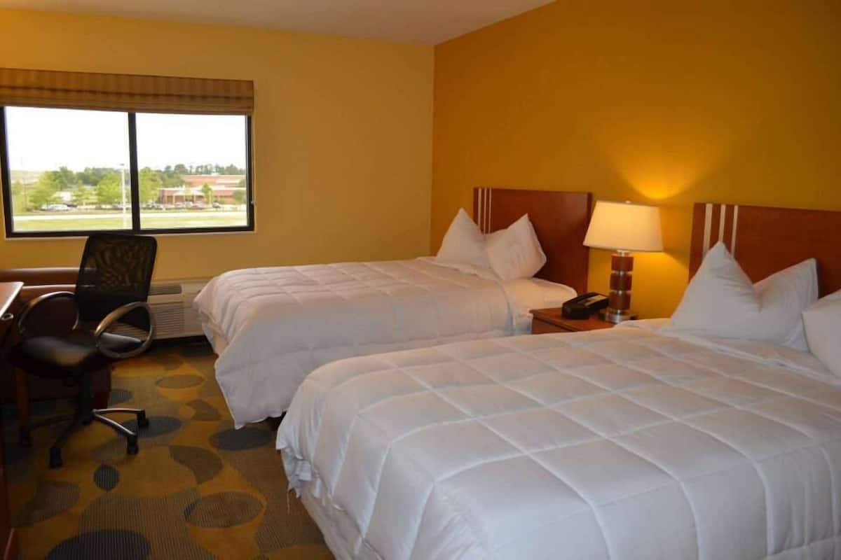 A hotel room with two beds, a desk, a chair, a lamp, and a window with a view of buildings and trees.