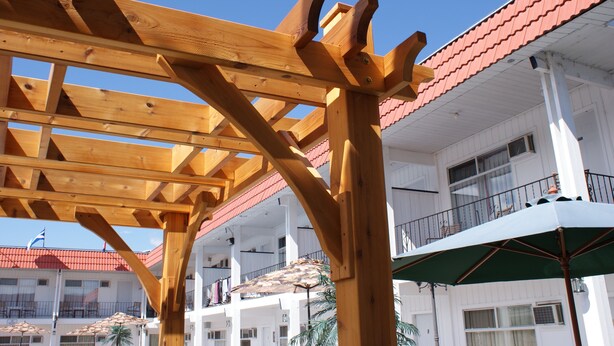 A wooden pergola structure with a red-tiled roof, white building with balconies, and a green umbrella. - Riverside Motel (Penticton)
