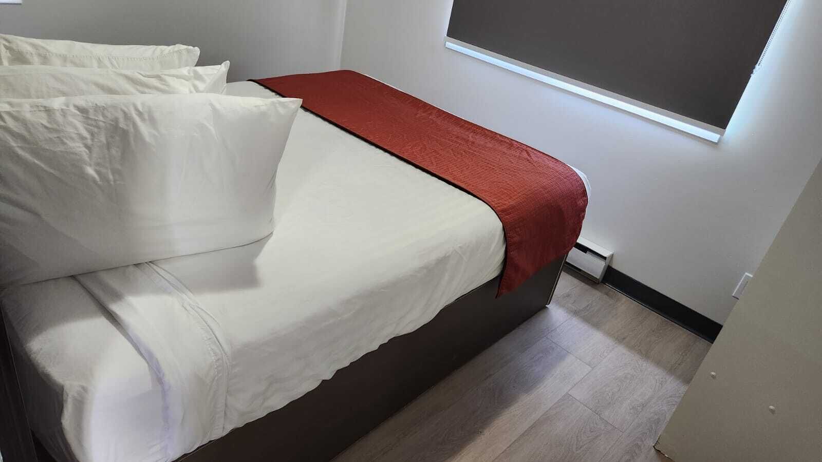 A bed with white linens and a red blanket, a window with blinds, and a light-colored wooden floor.