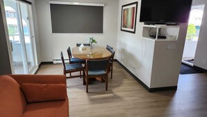 A modern living room with a round wooden dining table, blue cushioned chairs, a red sofa, a flat-screen TV mounted on the wall, and a small kitchenette area. - Riverside Motel (Penticton)