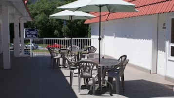 Pátio de motel com área de estar externa e guarda-sóis, um edifício branco com telhado vermelho e uma placa com o nome Riverside Motel.