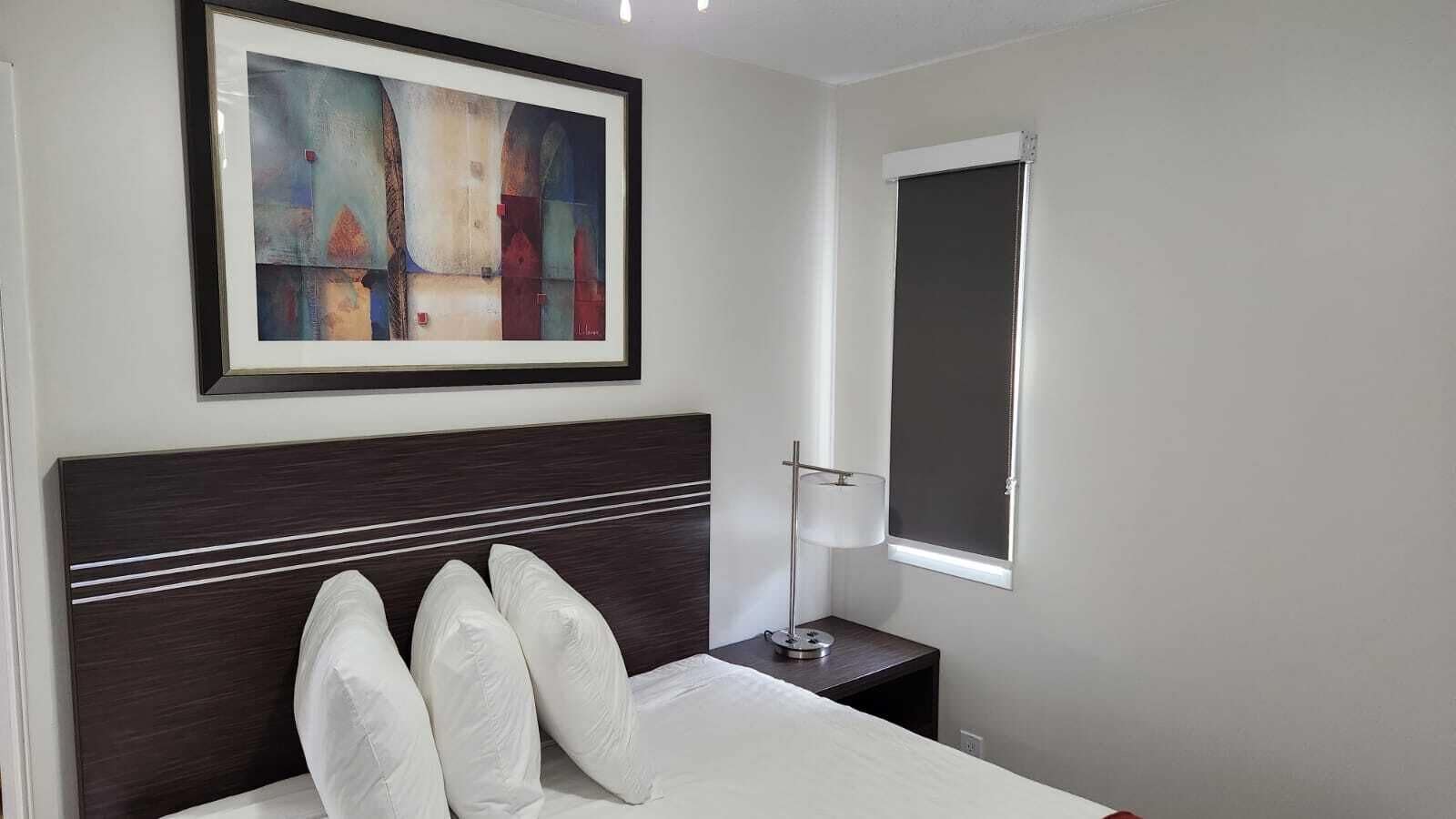 Habitación de hotel ordenada con cama, lámpara de noche, cuadro abstracto enmarcado y ventana con persiana.