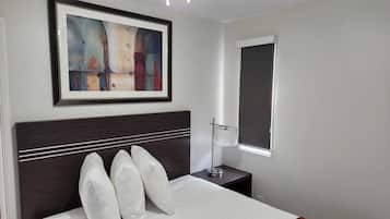 A neatly arranged hotel room with a bed, a bedside lamp, a framed abstract painting, and a window with a blind.