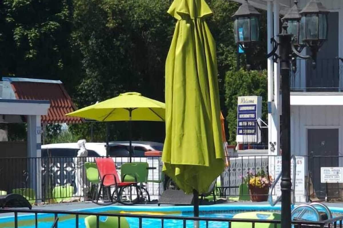 Uma área à beira da piscina com cadeiras e guarda-sóis verdes, um edifício vermelho com bandeiras e uma placa indicando um restaurante.