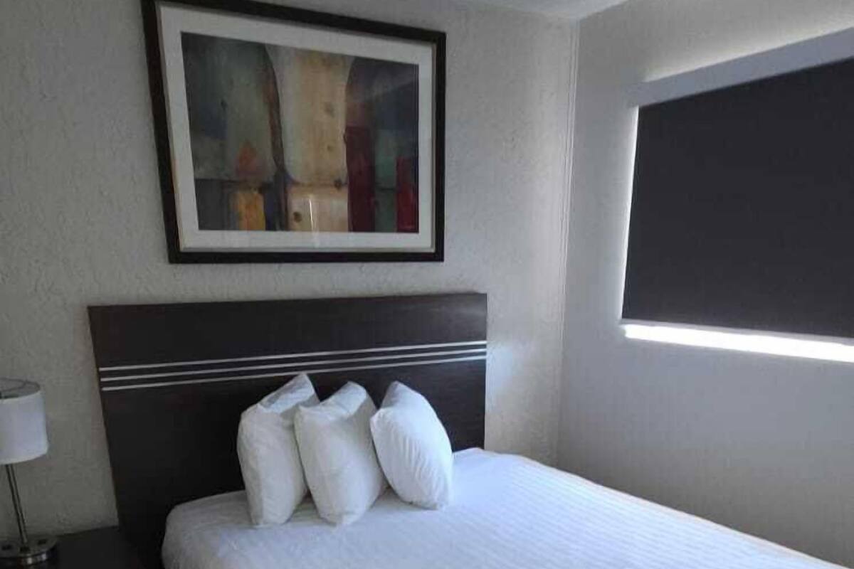 Quarto de hotel moderno com uma cama grande, uma mesa de cabeceira, uma obra de arte emoldurada na parede e uma janela com persianas.