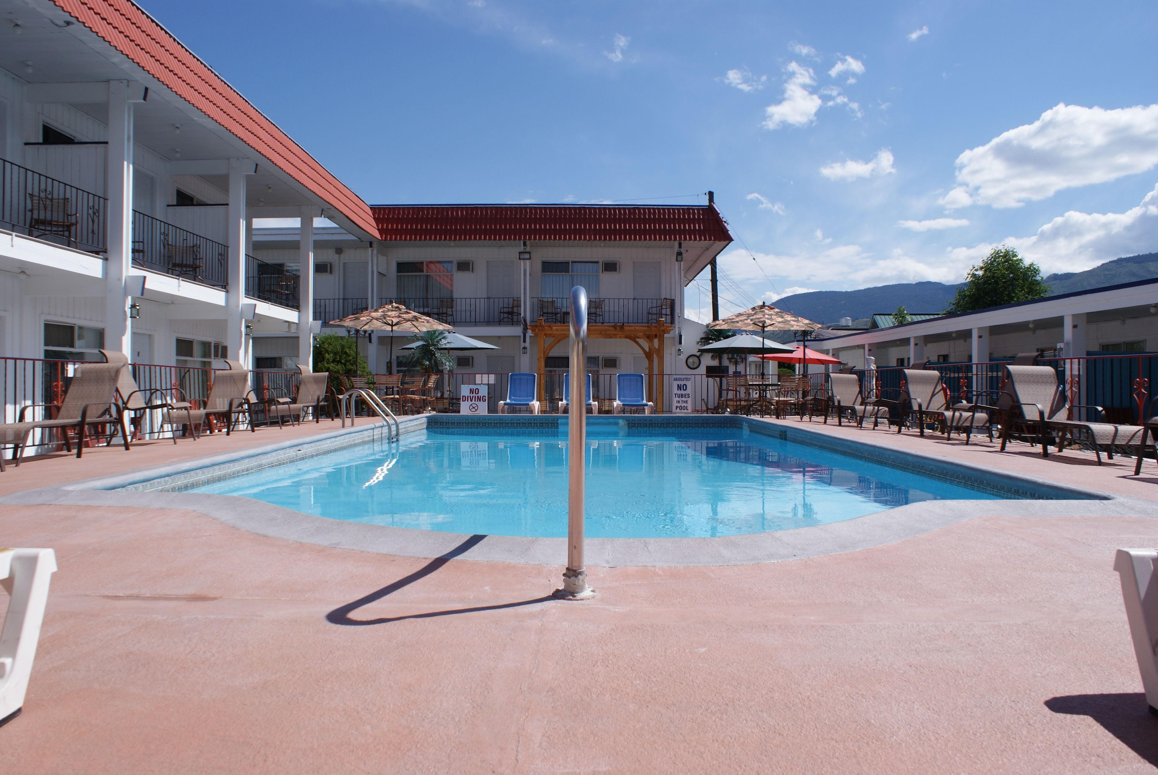 Ein Schwimmbad mit Liegestühlen und Sonnenschirmen, ein Gebäude mit Balkonen und eine gebirgige Kulisse.