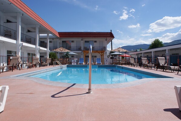 Una piscina con sillas de descanso y sombrillas, un edificio con balcones y un paisaje montañoso de fondo.