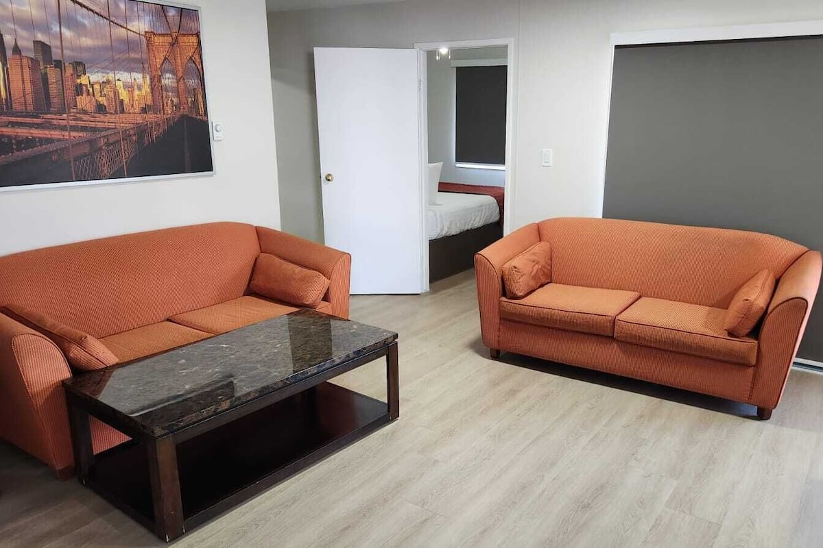 Uma sala de estar moderna com dois sofás laranja, uma mesa de centro de vidro e uma pintura com vista para a cidade na parede.