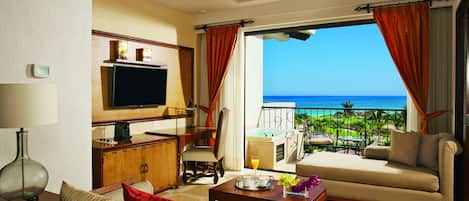 A hotel room with a balcony, a sofa, a TV, a coffee table, and a dining area with a view of the ocean.