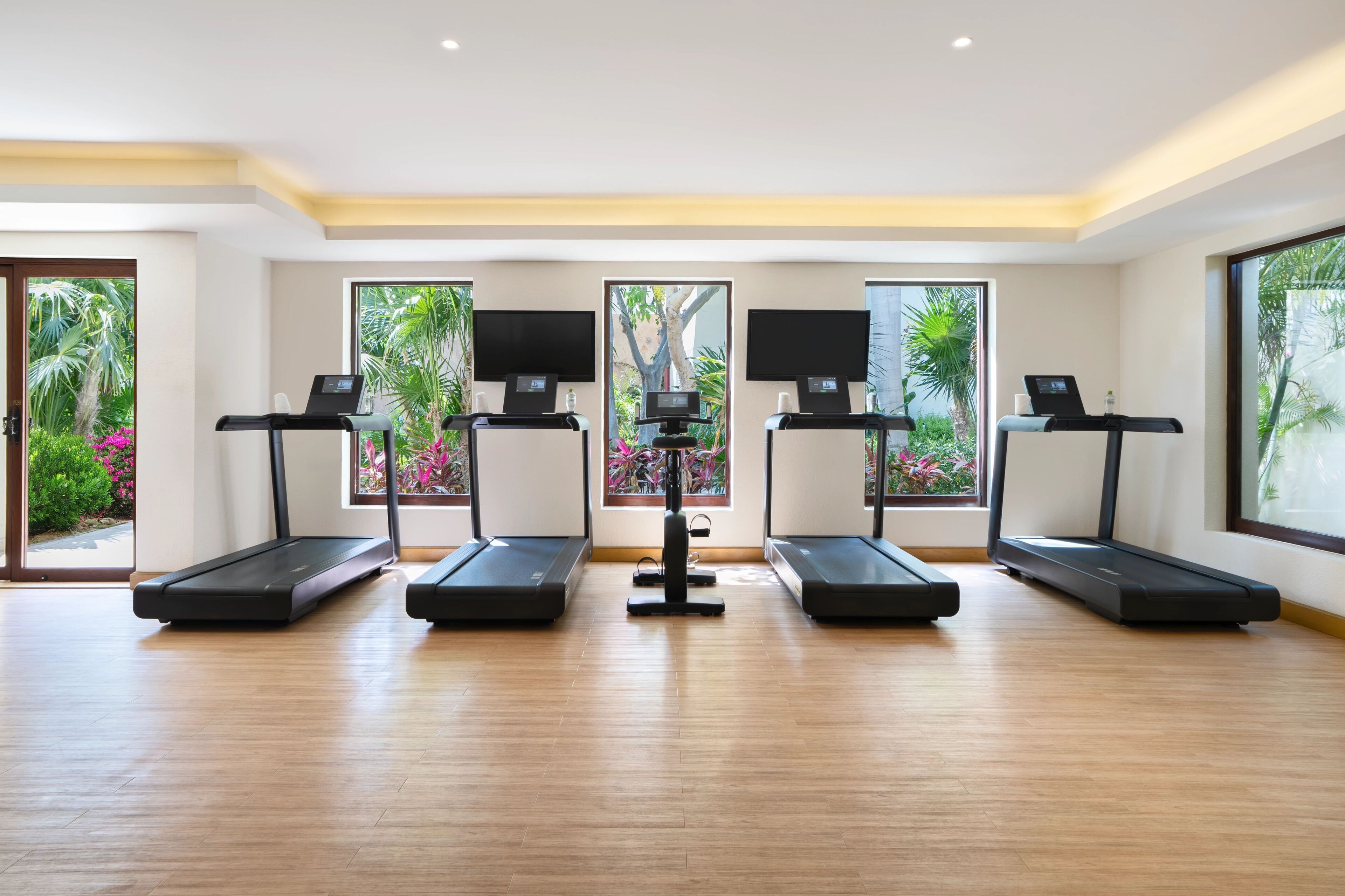 Un gimnasio con cuatro cintas de correr, una máquina de ejercicios central y grandes ventanales con vistas a zonas verdes.