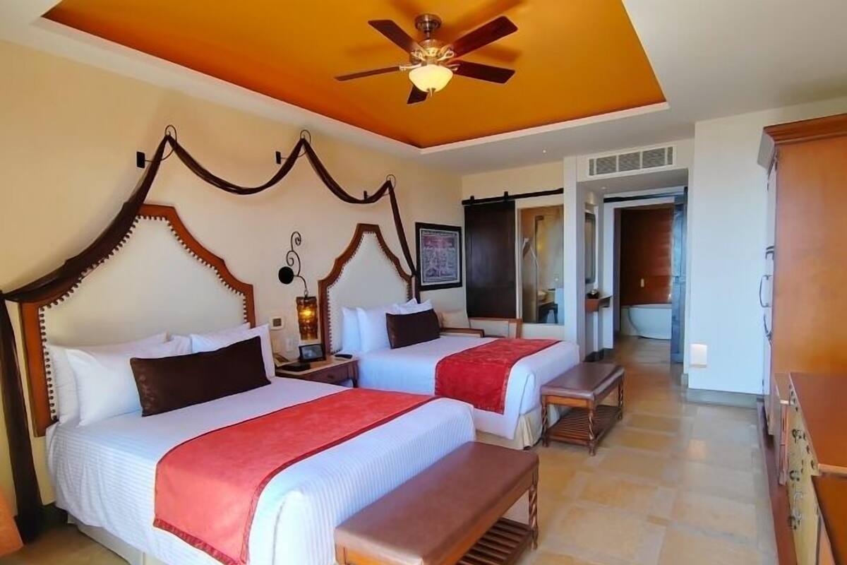 Quarto de hotel com duas camas, ventoinha no teto, um banco de madeira e banheiro visível por uma porta aberta.