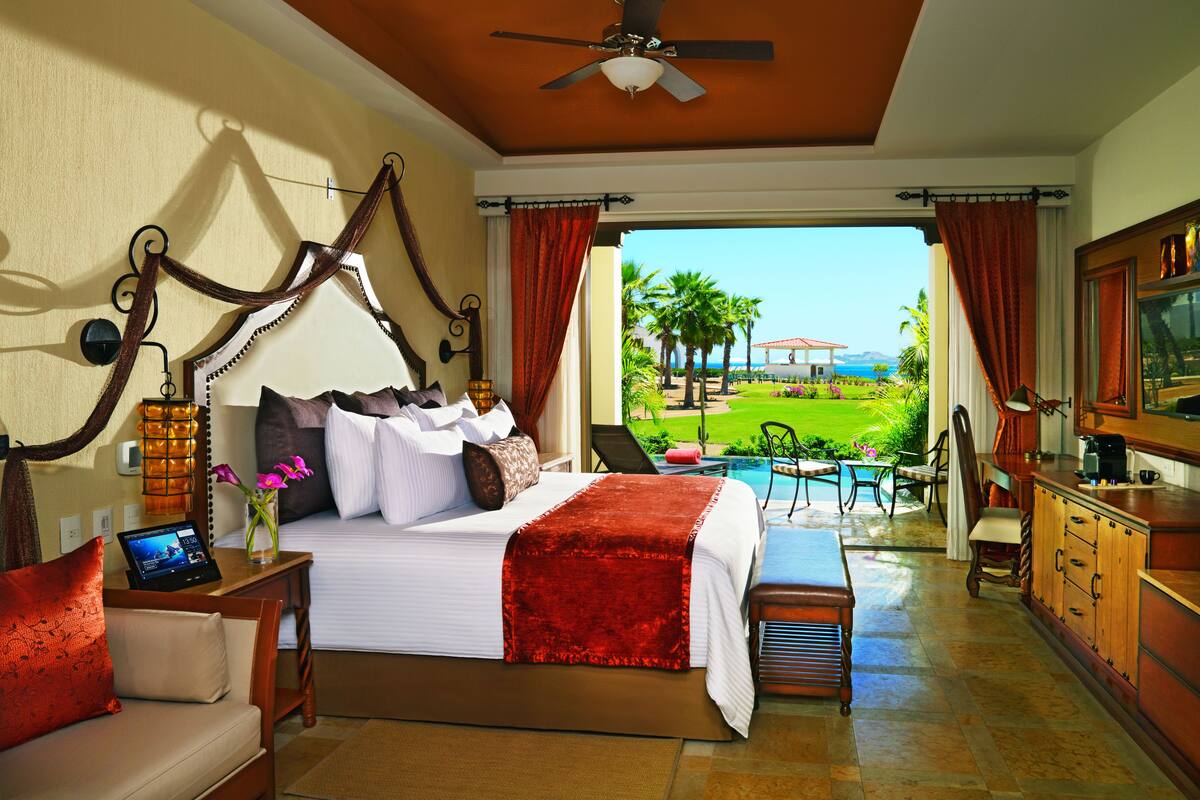Quarto de hotel com cama, escrivaninha, cadeira, televisão, janela com cortinas e vista para a piscina e jardim.