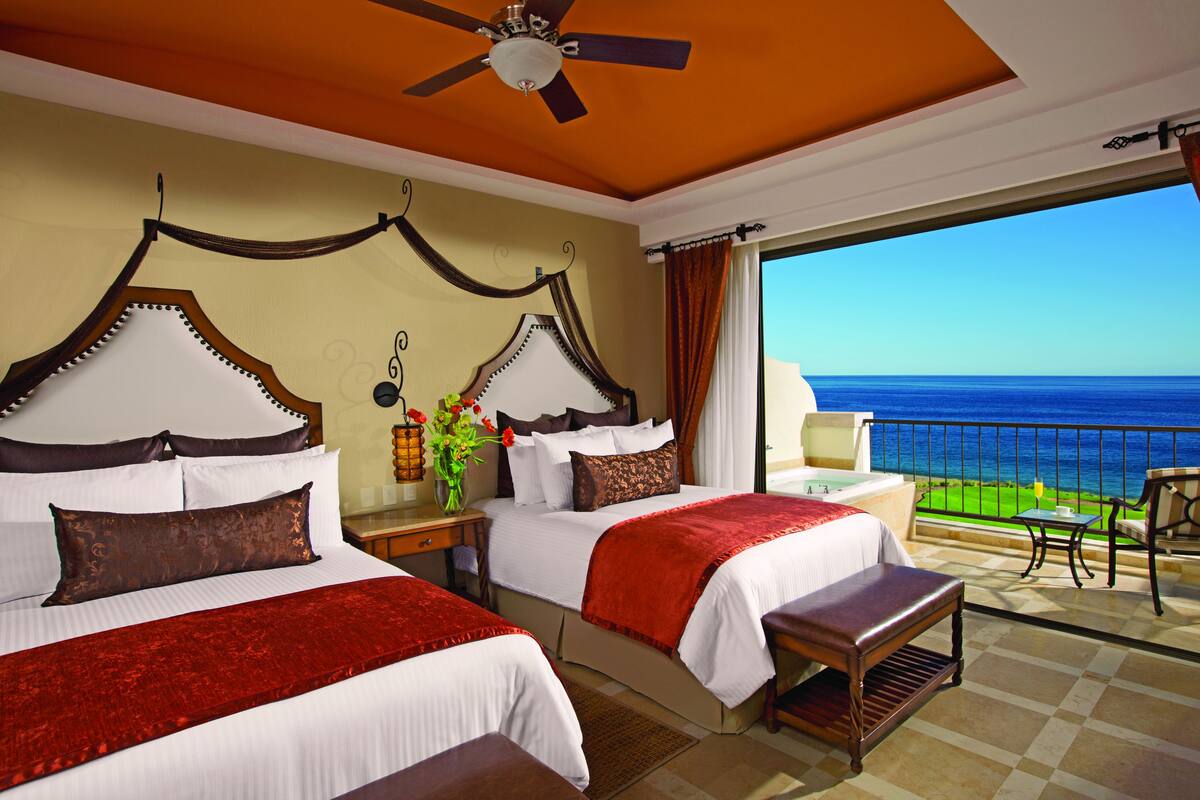 Quarto de hotel com duas camas, uma varanda com vista para o oceano, ventilador de teto e mesa de cabeceira com um vaso de flores.