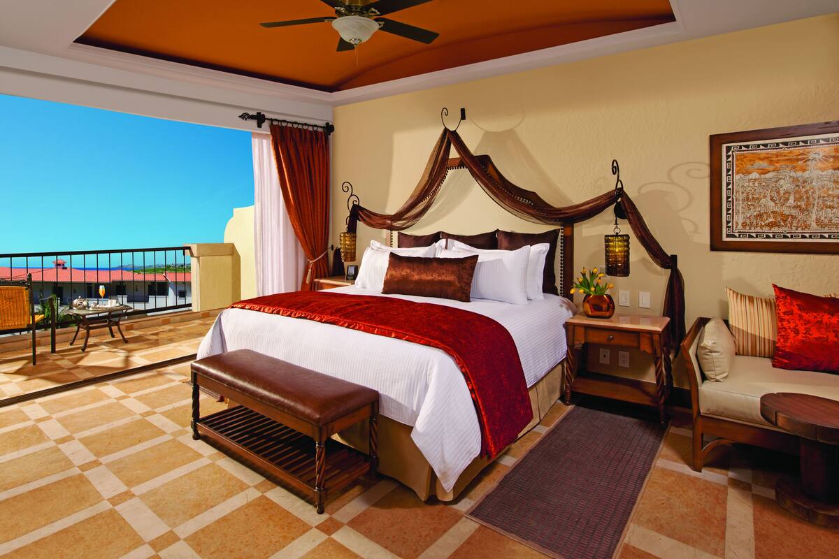 Um quarto espaçoso com uma cama grande, um banco de madeira, uma mesa de cabeceira e vista para o oceano.