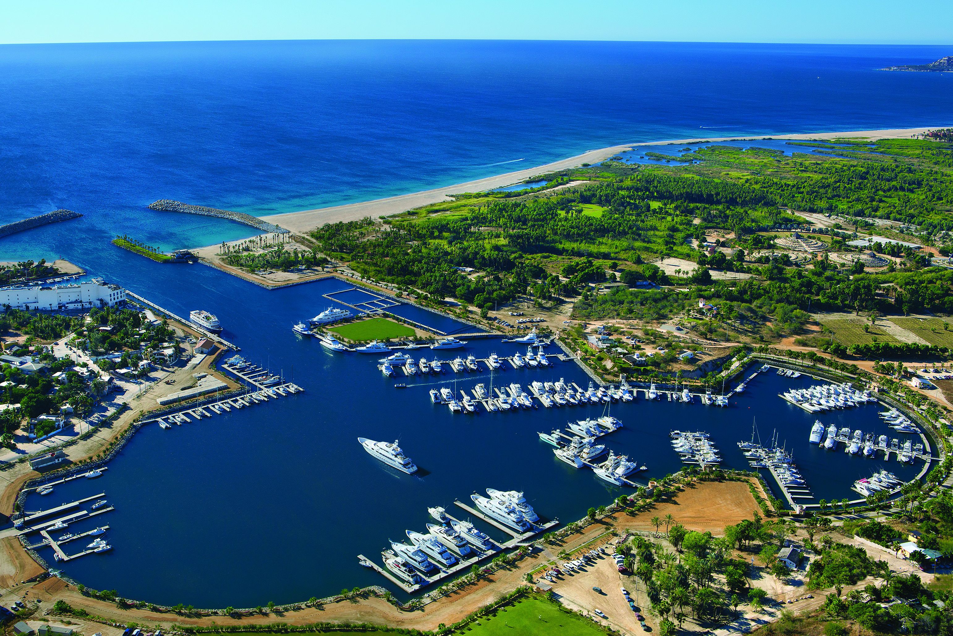 Vista aérea de un puerto deportivo con varios muelles y barcos, rodeado de vegetación y una playa.