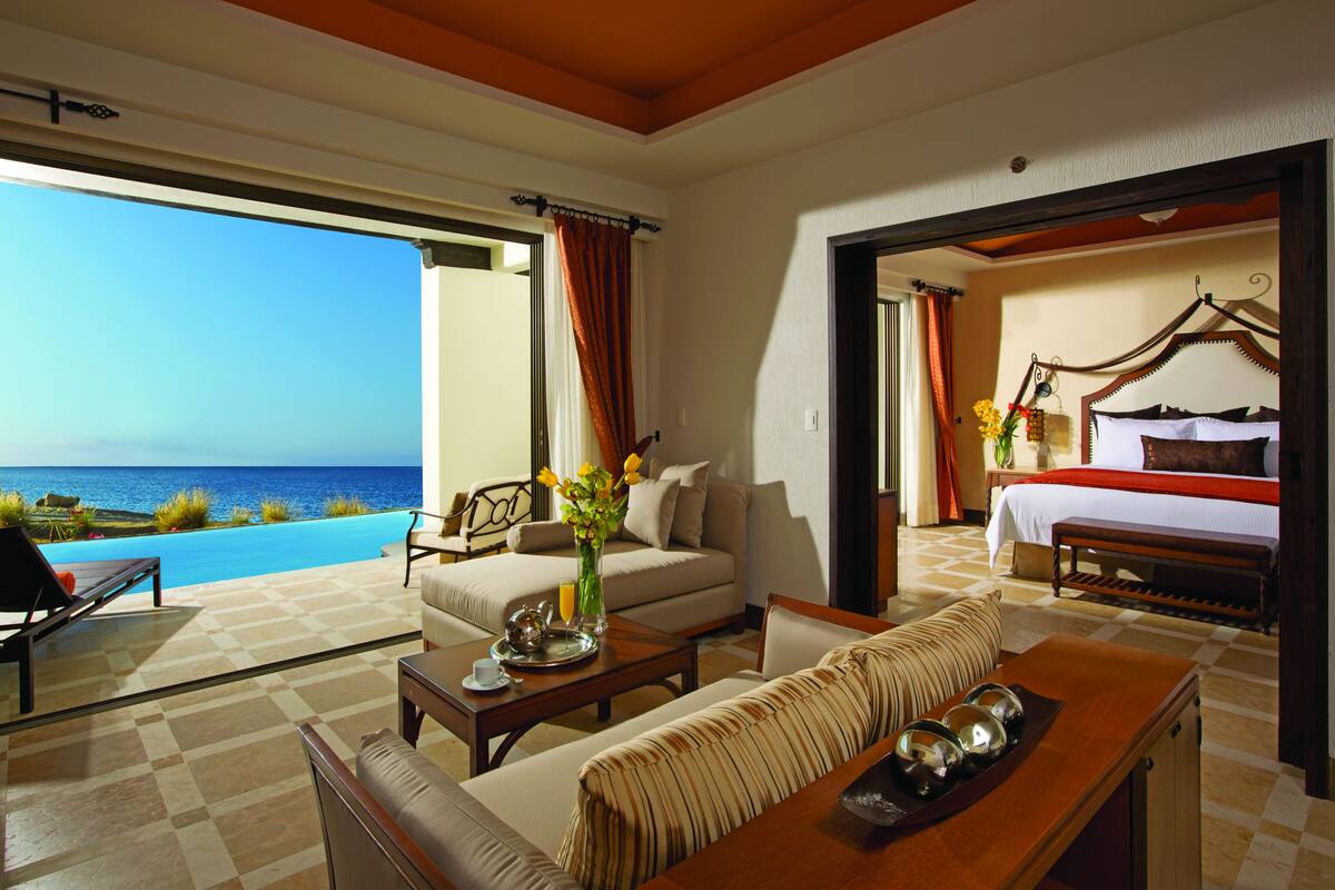 Quarto de hotel com uma janela ampla que dá para uma piscina e o mar, uma cama com cabeceira de madeira, um sofá e uma mesa de centro com um vaso de flores.