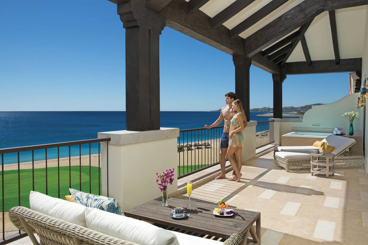 Uma área externa coberta com vista para o mar, uma mesa de jantar com uma travessa de frutas e uma bebida, e um espaço de estar com sofá e almofadas.