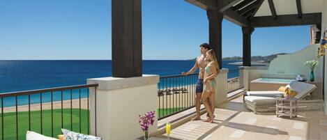 A covered outdoor area with a view of the sea, a dining table with a plate of fruit and a drink, and a lounge area with a sofa and cushions.