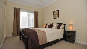 A bedroom with a bed, curtains, a ceiling fan, a bedside table with a lamp, and a wall-mounted picture.
