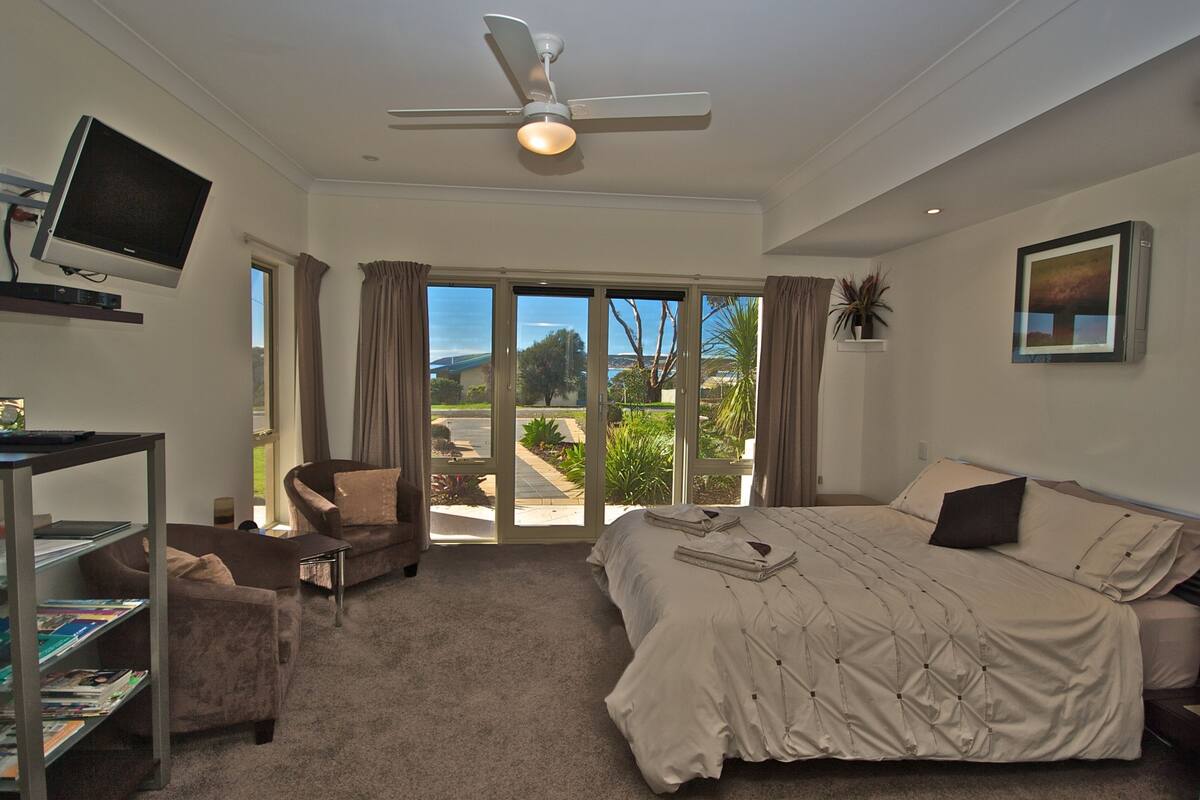 A bedroom with a bed, a desk with a TV, a chair, a bookshelf, and a view of the outdoors.