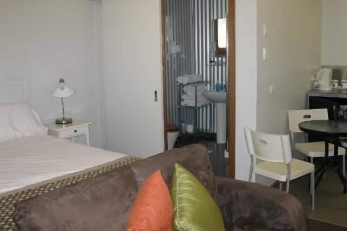 Hotellihuone, jossa on sänky, yöpöytä lampun kanssa, sohva tyynyineen, kylpyhuone suihkulla sekä ruokailutila tuoleineen ja pöytineen.