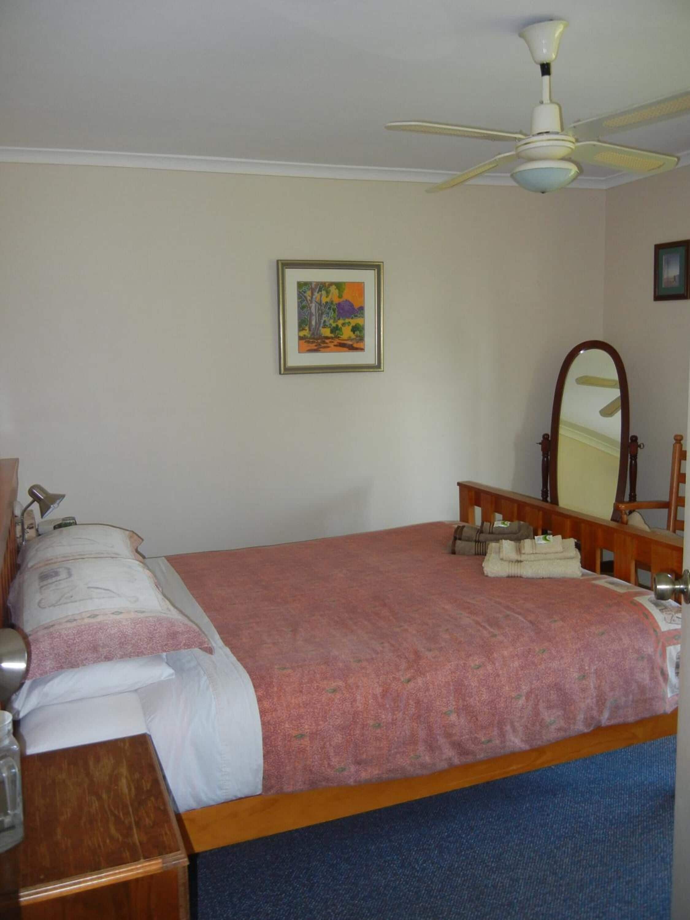 A bedroom with a bed, a ceiling fan, a bedside table, a lamp, a mirror, and a framed picture on the wall.
