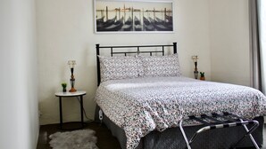 A bedroom with a bed, a small bedside table with a lamp, a potted plant, and a painting on the wall. - The Apartment (Moyston)