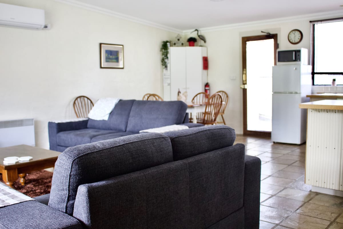 A living room with a grey sofa, a dining table with chairs, a kitchen area with a microwave, and a white cabinet.