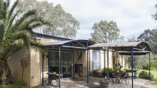 A covered patio area with a table and chairs, a pergola, and a pathway leading to a glass door entrance. - The Apartment (Moyston)