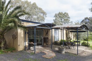 A covered patio area with a table and chairs, a pergola, and a pathway leading to a glass door entrance. - The Apartment (Moyston)