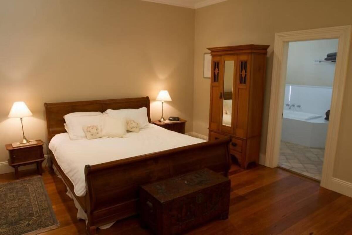 A bedroom with a wooden bed, two bedside lamps, a wooden nightstand, a wooden chest of drawers, and a view of a bathroom through an open door.