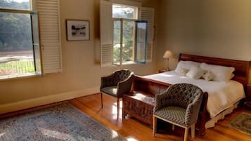 A bedroom with a bed, two chairs, a nightstand, a window with shutters, and a view of greenery outside.
