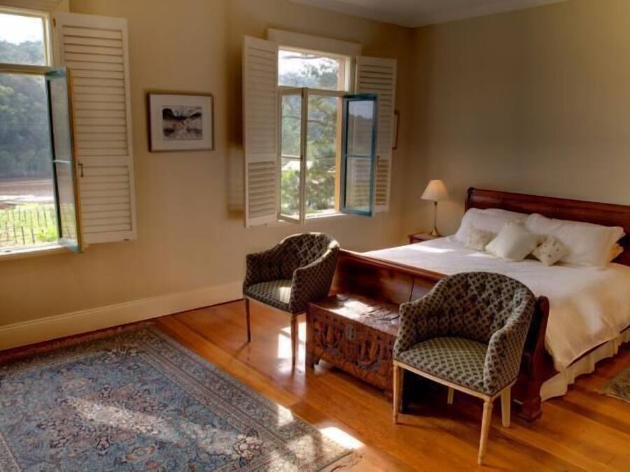 Une chambre à coucher comprenant un lit, deux chaises, une table de chevet, une fenêtre avec des volets et une vue sur la verdure à l’extérieur.