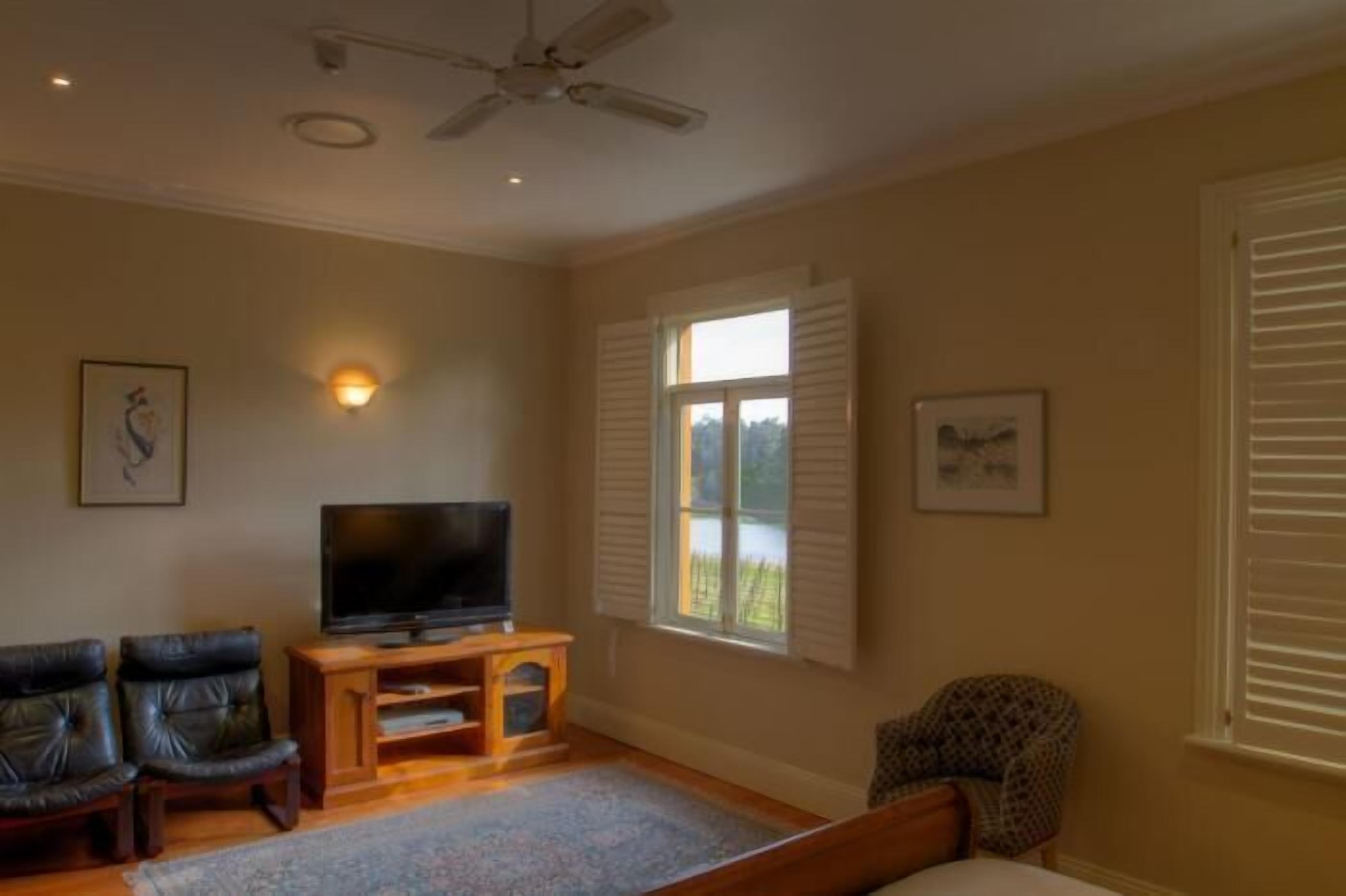 Un salon avec un téléviseur à écran plat posé sur un meuble en bois, deux fauteuils en cuir, un fauteuil à motifs, un ventilateur de plafond et une fenêtre avec des volets.