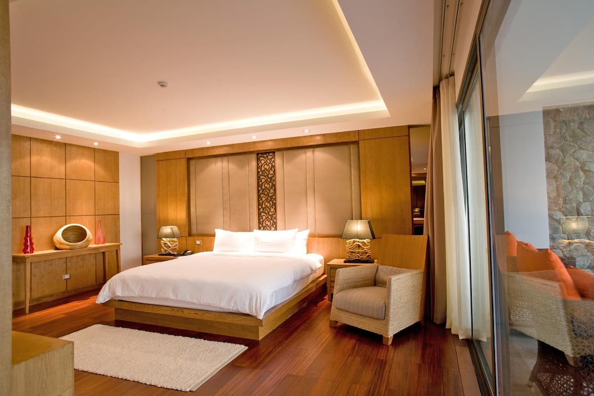 A modern bedroom with a large bed, wooden paneling, a glass door leading to a balcony, and a small seating area.